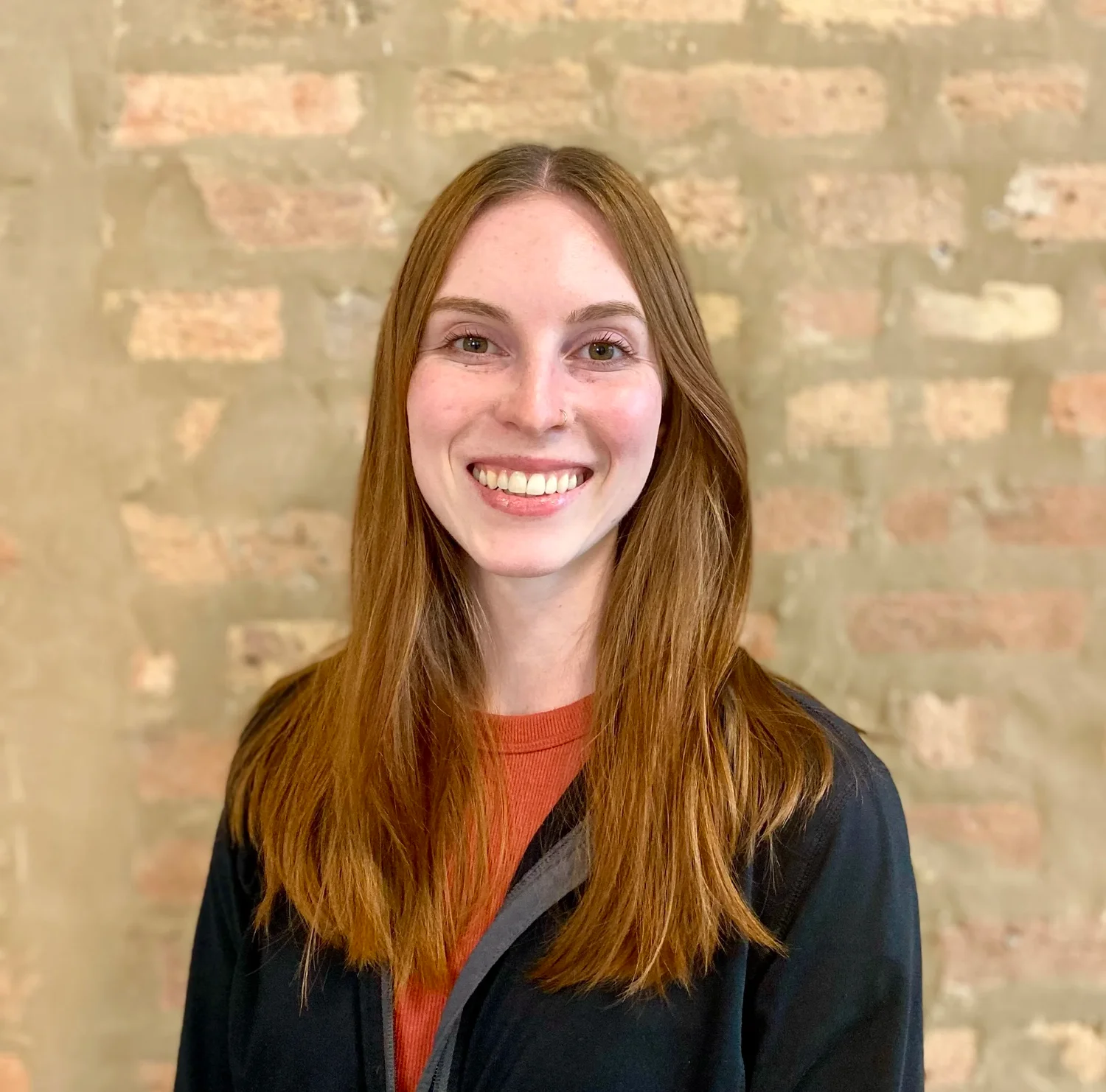 Rachel Reynolds, smiling physical therapist with long red hair, wearing a black jacket over an orange top, against a textured brick wall.